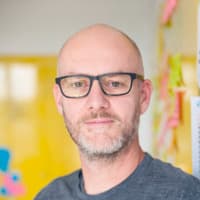 A bald man with a three day, slightly grey beard and black glasses looking into the camera.