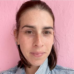 A woman with brown shoulder-length hair, brown eyes and freckles looking into the camera in front of a pink background.