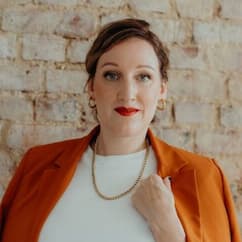 A woman in a bright orange blazer and white shirt with brown hair in a bun looking into the camera.